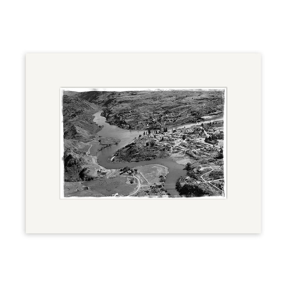 The Alexandra 1956 - HI102 is a matte black-and-white aerial photo featuring a winding river, rocky hills, buildings, roads, and bridges. This historic print comes framed with a wide white mat for archival-quality display.