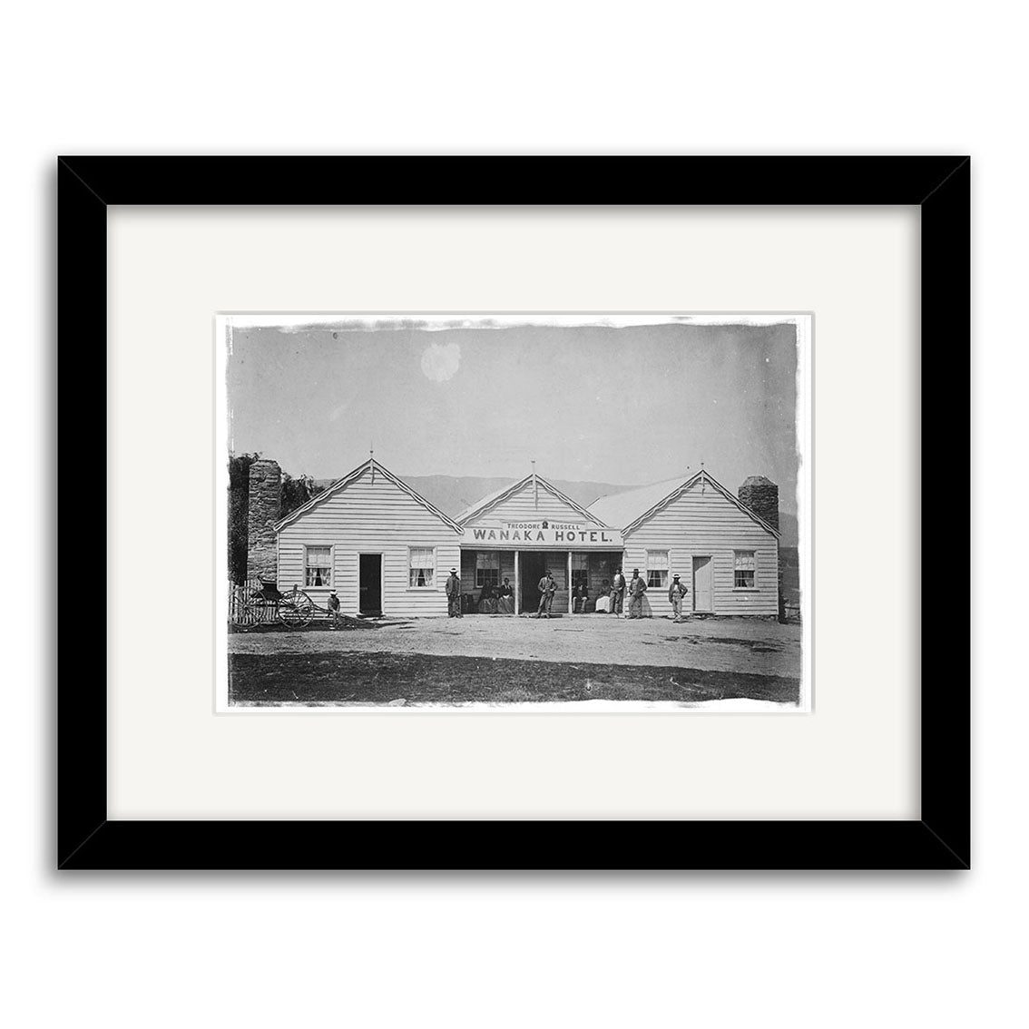 A black-and-white framed historical print titled Wanaka Hotel 1870 - HI104, showing people outside an old wooden building in New Zealand with bicycles nearby. Printed on archival quality matte paper.