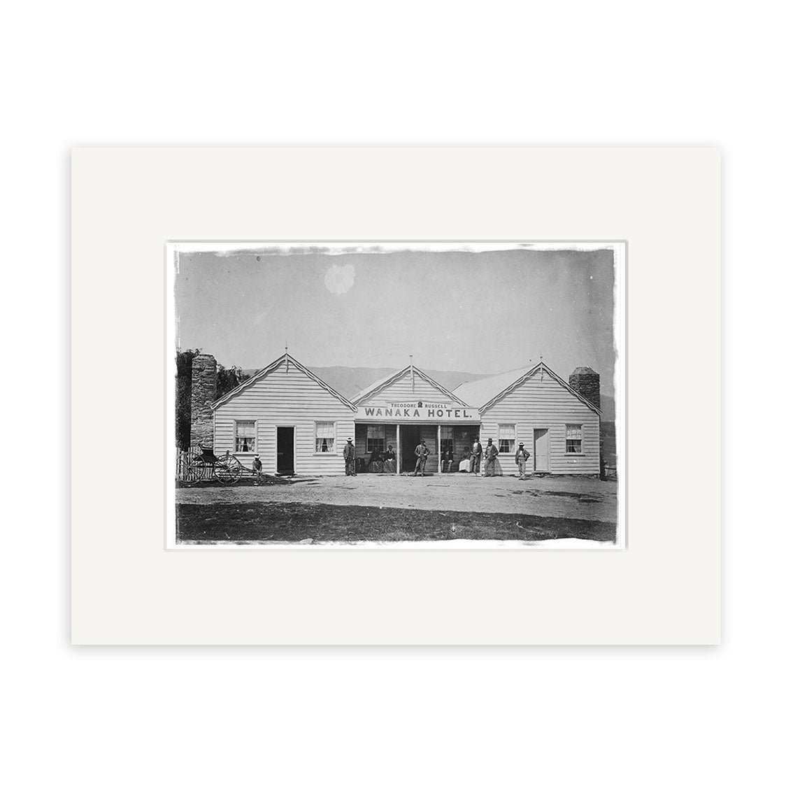 Black-and-white photo of Wanaka Hotel 1870 - HI104, a single-story wooden building in New Zealand, with people out front, horse and carriage on the left, and mountains behind—archival quality matte paper Historical Photographic Print.