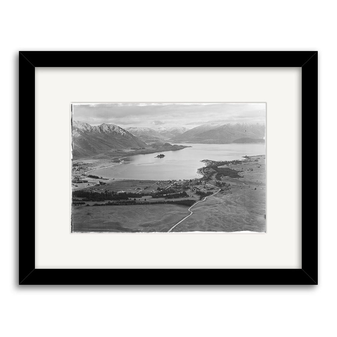 Lake Wanaka 1951 - HI105: Archival quality black and white framed print of a wide lake in New Zealand, surrounded by mountains, open land, a small island in the center, and a curving road in the foreground.