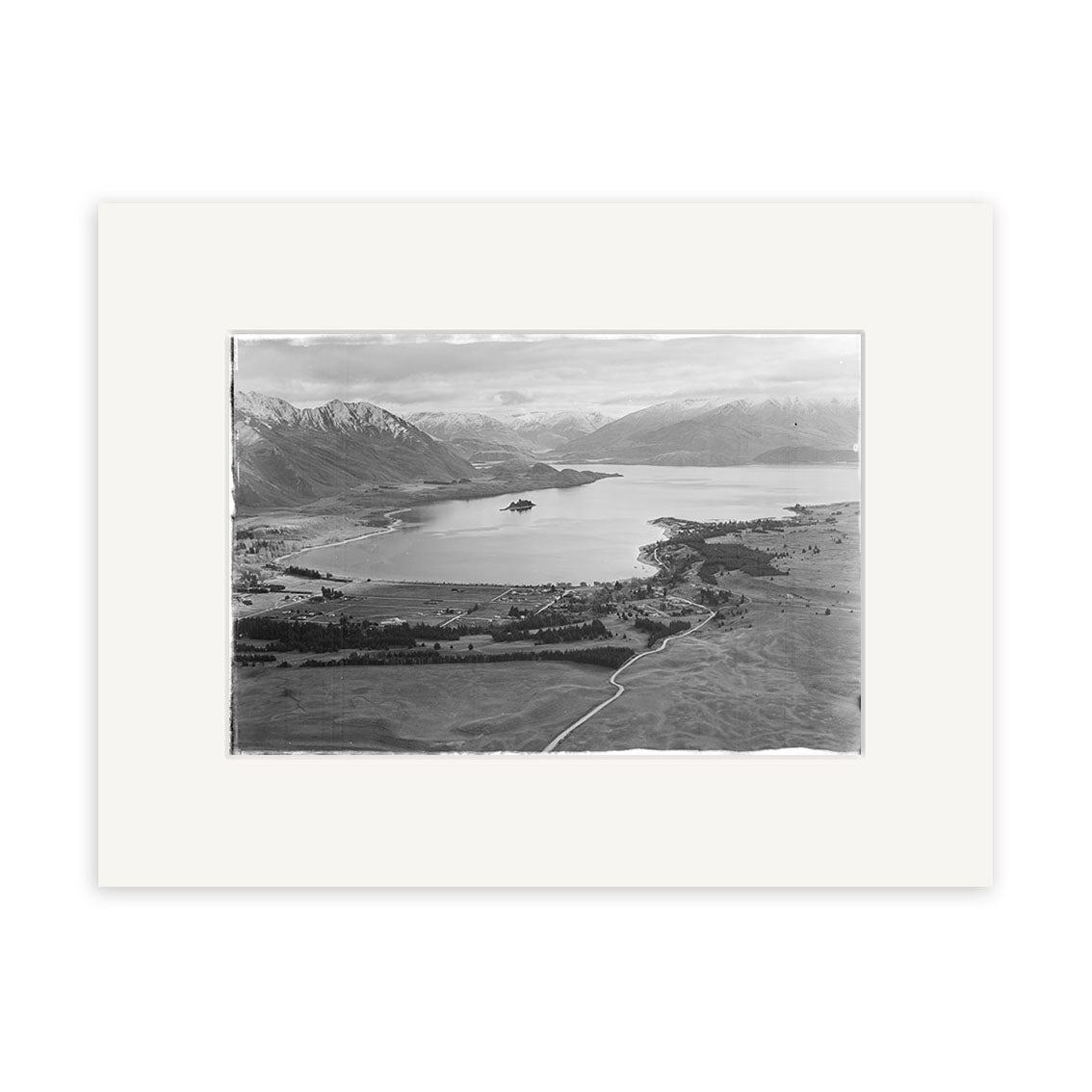 A black and white aerial photo, Lake Wanaka 1951 - HI105, shows a wide lake surrounded by fields and mountains in New Zealand. Roads and small buildings appear near the shore. This historical photographic print is centered on a white mat.