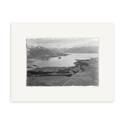 A black and white aerial photo, Lake Wanaka 1951 - HI105, shows a wide lake surrounded by fields and mountains in New Zealand. Roads and small buildings appear near the shore. This historical photographic print is centered on a white mat.