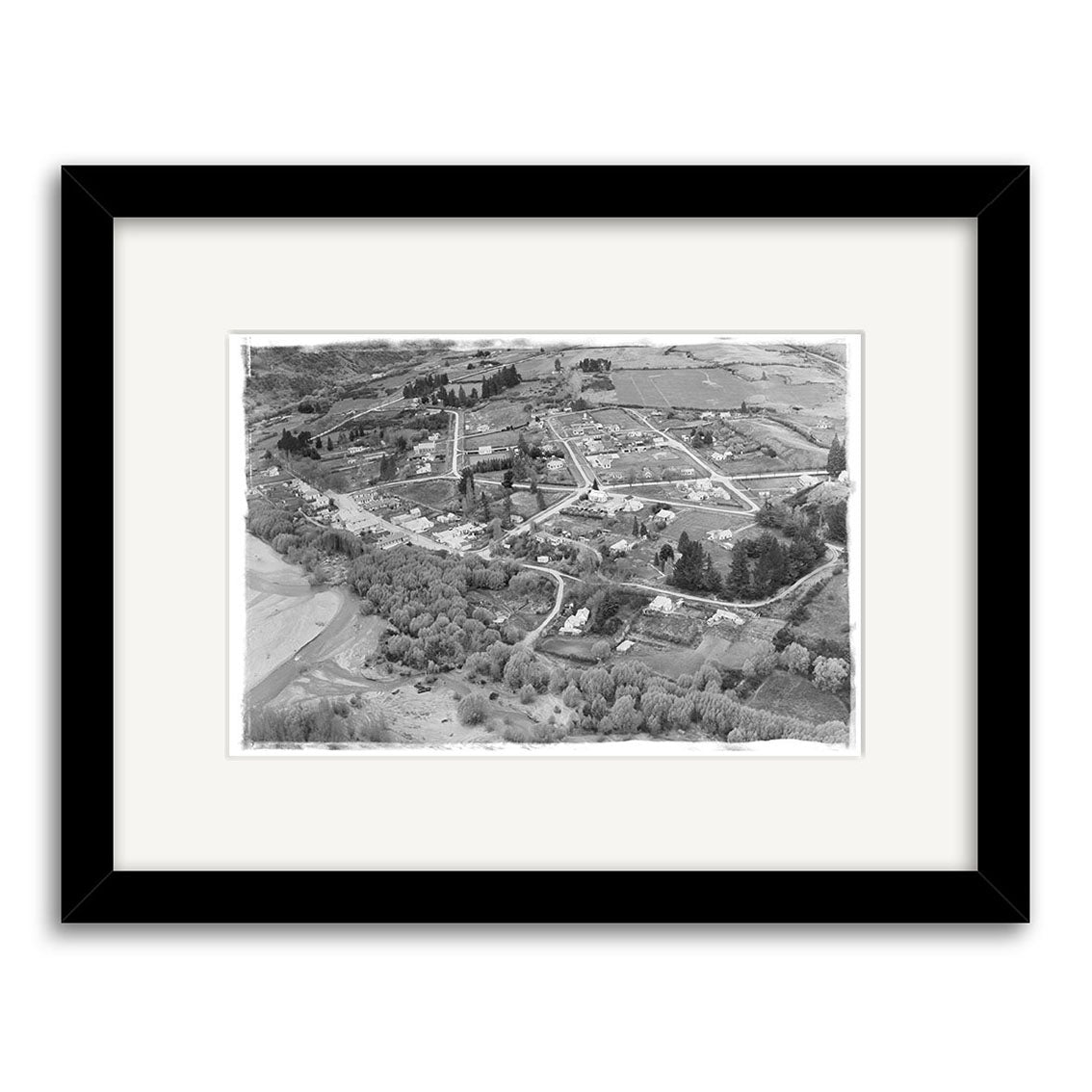 Aerial black and white photograph titled Arrowtown 1951 - HI106, showing a rural New Zealand area with scattered buildings, grid roads, open fields, and trees. Presented on archival matte paper, framed with a white mat and black frame.