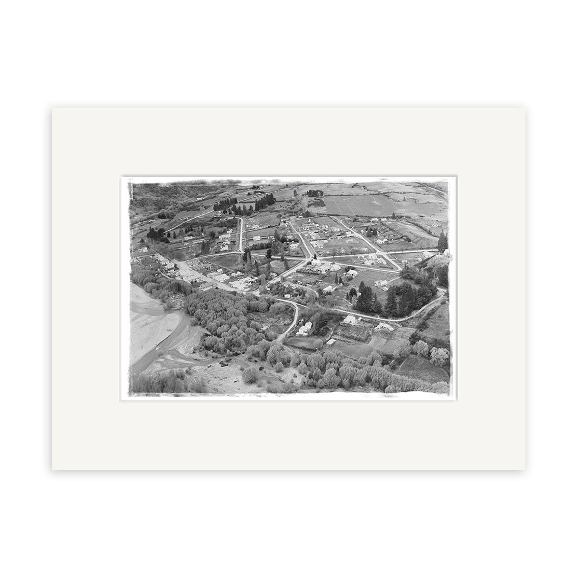 Arrowtown 1951 - HI106 is a black-and-white aerial photo of a small New Zealand town, depicting streets, buildings, fields, and trees. This historic print is on archival quality matte paper with a white border.