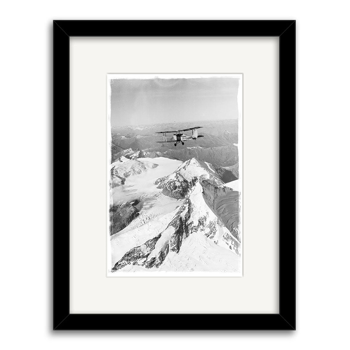 Mt Aspiring 1950 - HI107: Black and white photo of a biplane above snowy peaks, viewed from behind. Framed with a simple black and white mat, this archival-quality print ensures lasting beauty.