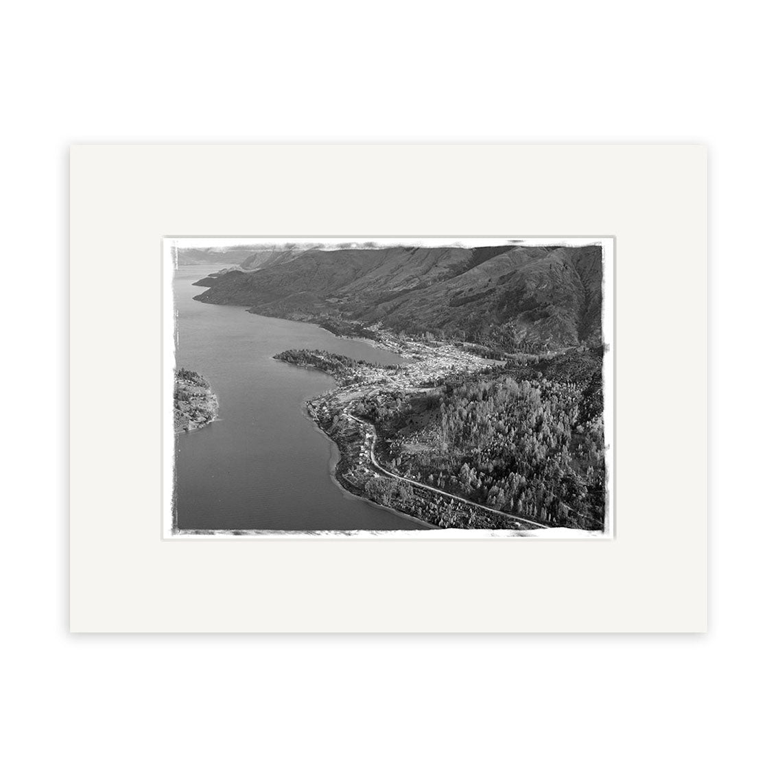 The Queenstown 1957 - HI108 is a black and white aerial photograph, printed on archival quality paper with a white border, showing the town between a lake and forested hills. Made in New Zealand.