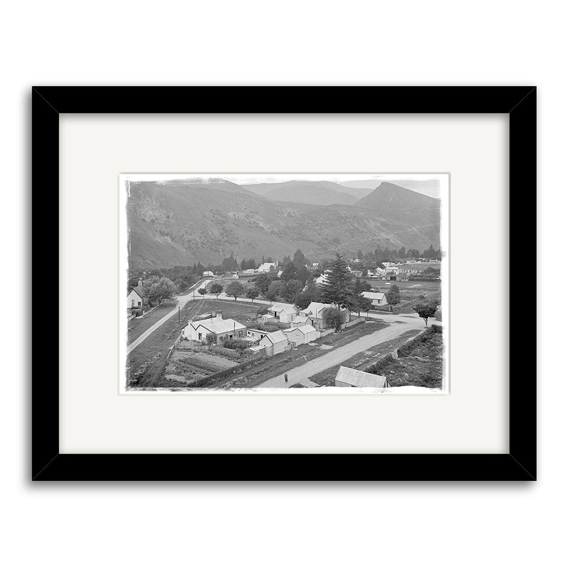 Arrowtown 1926 - HI109: Black and white historical photo print of a rural town with houses, wide road, trees, and hills; displayed in a black frame with white mat. Made in New Zealand with archival quality.