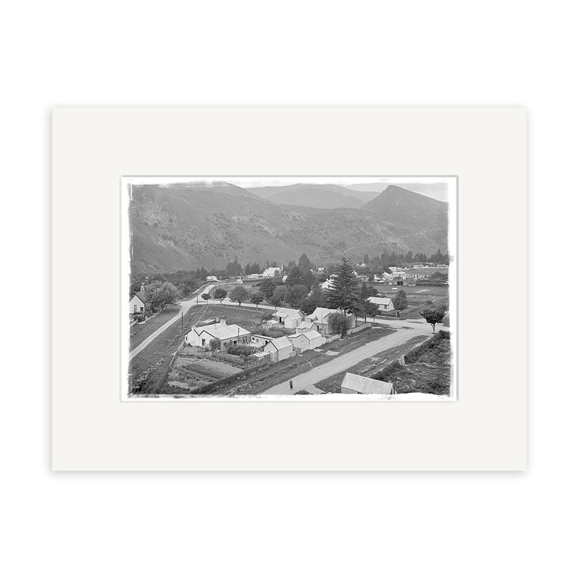 A black and white photograph titled Arrowtown 1926 - HI109 shows a rural New Zealand town with houses, roads, trees, and mountains. This historical print is framed with a wide white mat.