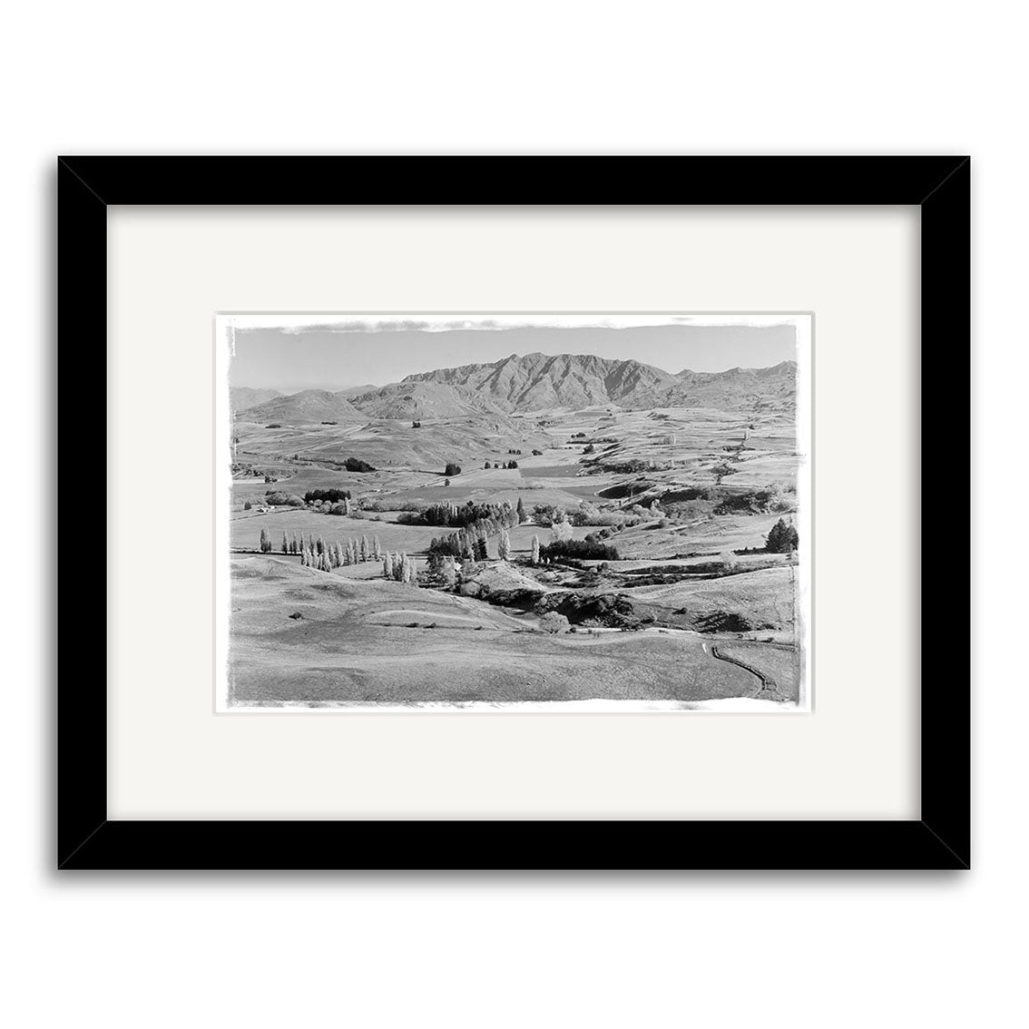 Black and white photographic print, Arrowtown District 1954 - HI111, features a rural New Zealand landscape with rolling hills, scattered trees, and distant mountains. Presented in a black frame with a white mat.