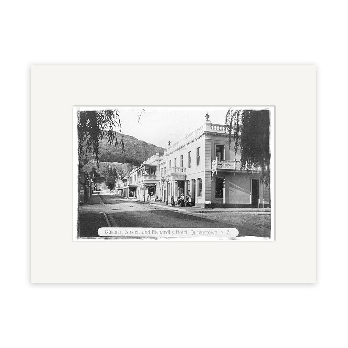 Black-and-white historical print Balarat St 1890 - HI112 shows Ballarat Street, Queenstown, NZ, with people outside Eichardt’s Hotel and buildings, trees, and hills visible in the background. Archival quality image.