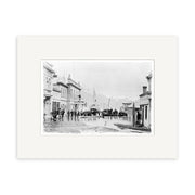 Black and white historical photographic print, Ballarat St Queenstown 1878 - HI113, shows people on a rain-wet street lined with buildings in New Zealand, with a boat and mountains in the background. Matted with a white archival-quality border.