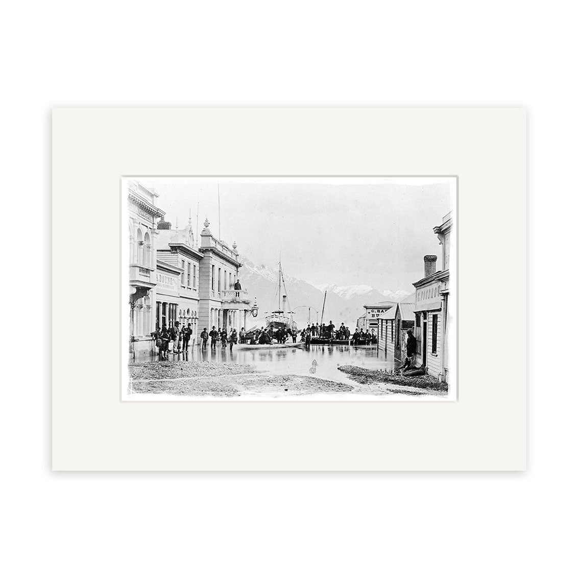 Black and white historical photographic print, Ballarat St Queenstown 1878 - HI113, shows people on a rain-wet street lined with buildings in New Zealand, with a boat and mountains in the background. Matted with a white archival-quality border.