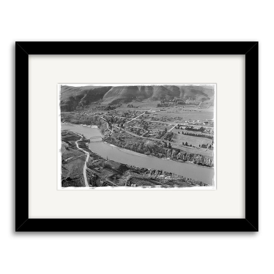 The Clyde 1957 - HI114 is a black-and-white aerial photograph of a river winding through rural New Zealand with a bridge, fields, and roads. This historical print comes in a simple black frame with a white mat for archival display.