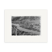 The product Clyde 1957 - HI114 is a black and white aerial photo showing a river, bridge, trees, fields, and buildings in New Zealand. This historic photographic print comes mounted on an archival quality white mat.