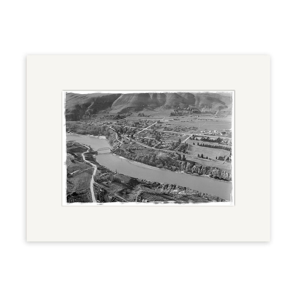The product Clyde 1957 - HI114 is a black and white aerial photo showing a river, bridge, trees, fields, and buildings in New Zealand. This historic photographic print comes mounted on an archival quality white mat.
