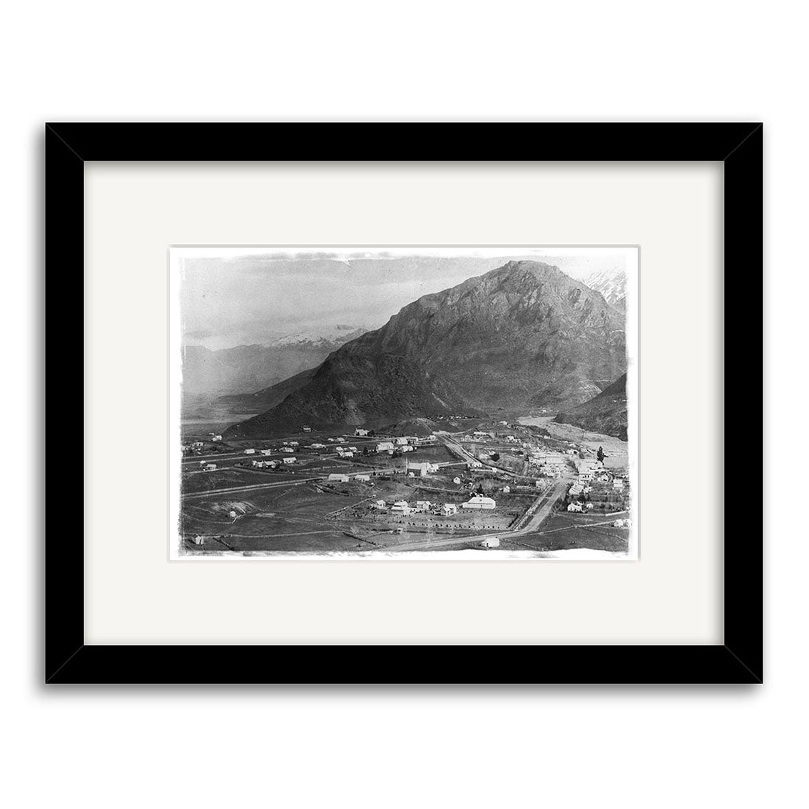 Arrowtown 1880 - HI116 is a black-and-white photo print of a small town with scattered buildings at the base of a large mountain, presented in a black frame with white mat.