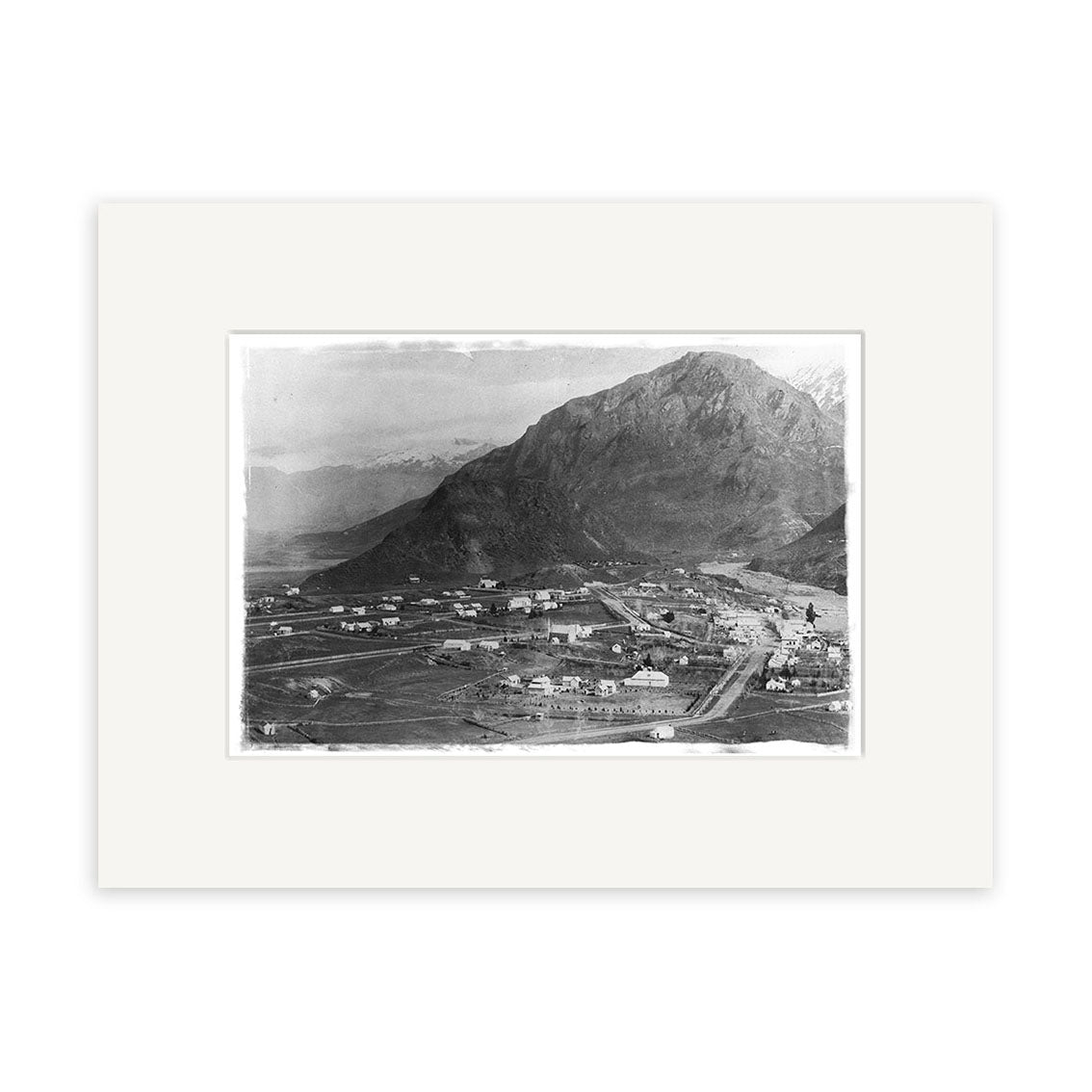Arrowtown 1880 - HI116: A black and white historical photographic print of a small town and mountain on archival matte paper in a sleek black frame, featuring roads, fields, scattered buildings, and distant snow-capped peaks.