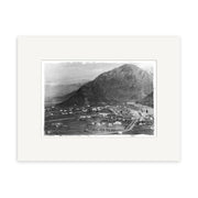 Arrowtown 1880 - HI116: A black and white historical photographic print of a small town and mountain on archival matte paper in a sleek black frame, featuring roads, fields, scattered buildings, and distant snow-capped peaks.