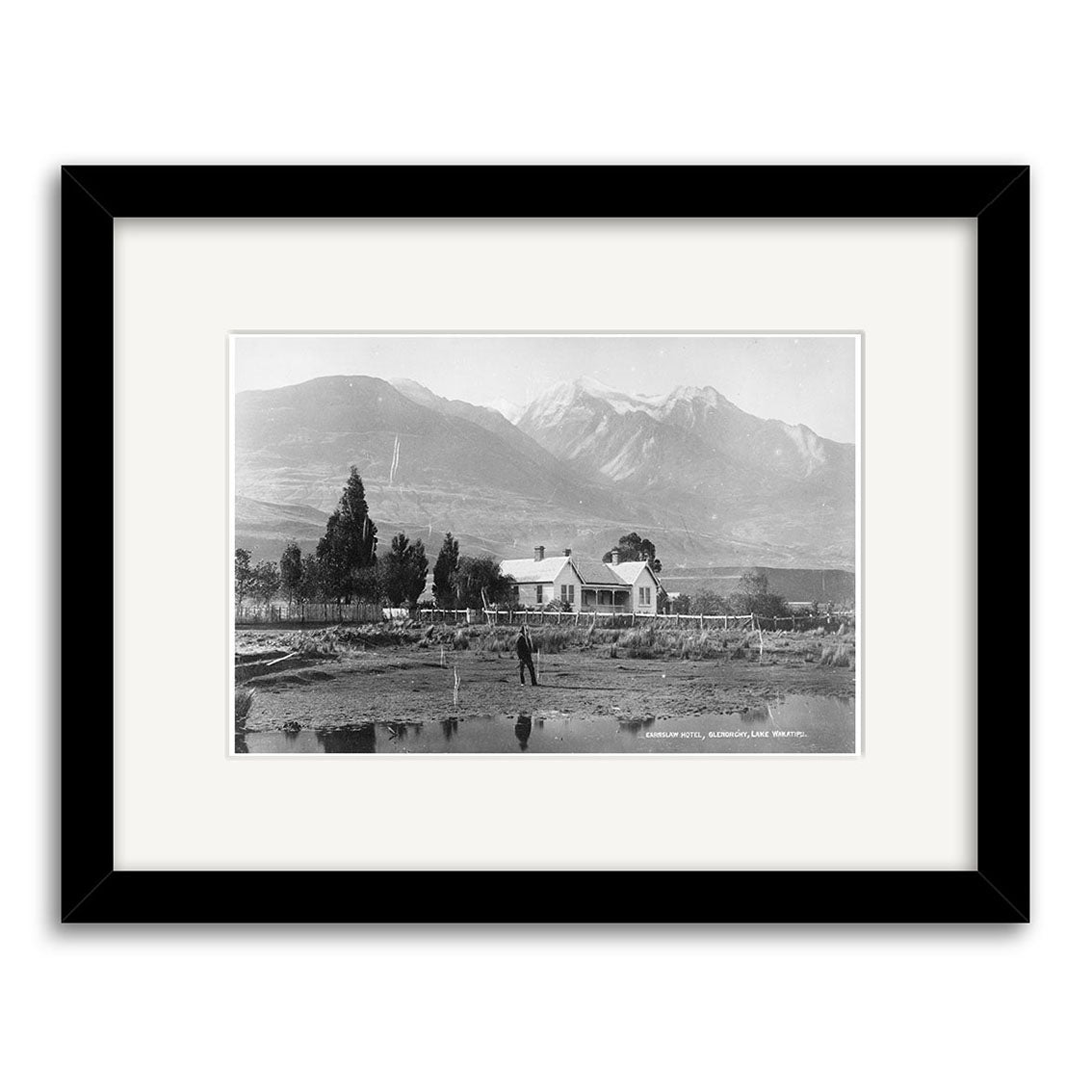 Earnslaw Hotel 1880 - HI117: Archival black and white photo print of a house with picket fence, trees, a person nearby, and mountains. Comes framed with a wide white mat and black frame.
