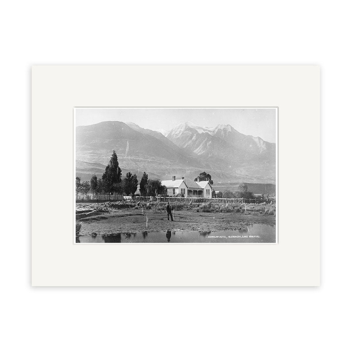 Archival quality black and white photo print, Earnslaw Hotel 1880 - HI117, shows a house with fenced yard, trees, and a person in front. Mountains rise in the background; includes classic white mat border.