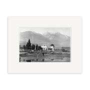Archival quality black and white photo print, Earnslaw Hotel 1880 - HI117, shows a house with fenced yard, trees, and a person in front. Mountains rise in the background; includes classic white mat border.