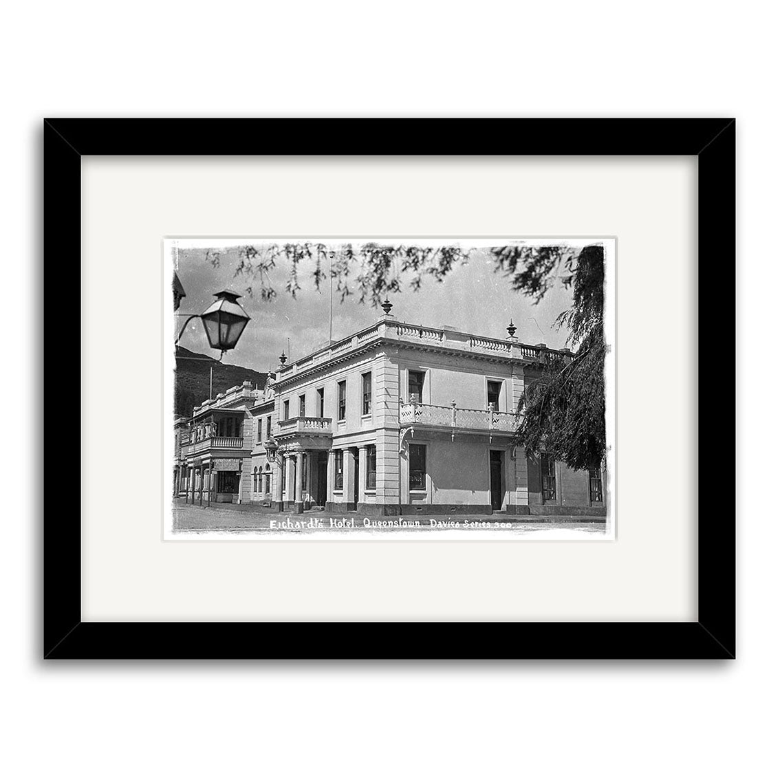 Black and white historical photographic print of Eichardts Hotel 1910 (HI118) in Queensland, featuring a two-story corner building with balconies and ornate railings amid other historic buildings and a streetlamp. Archival quality.