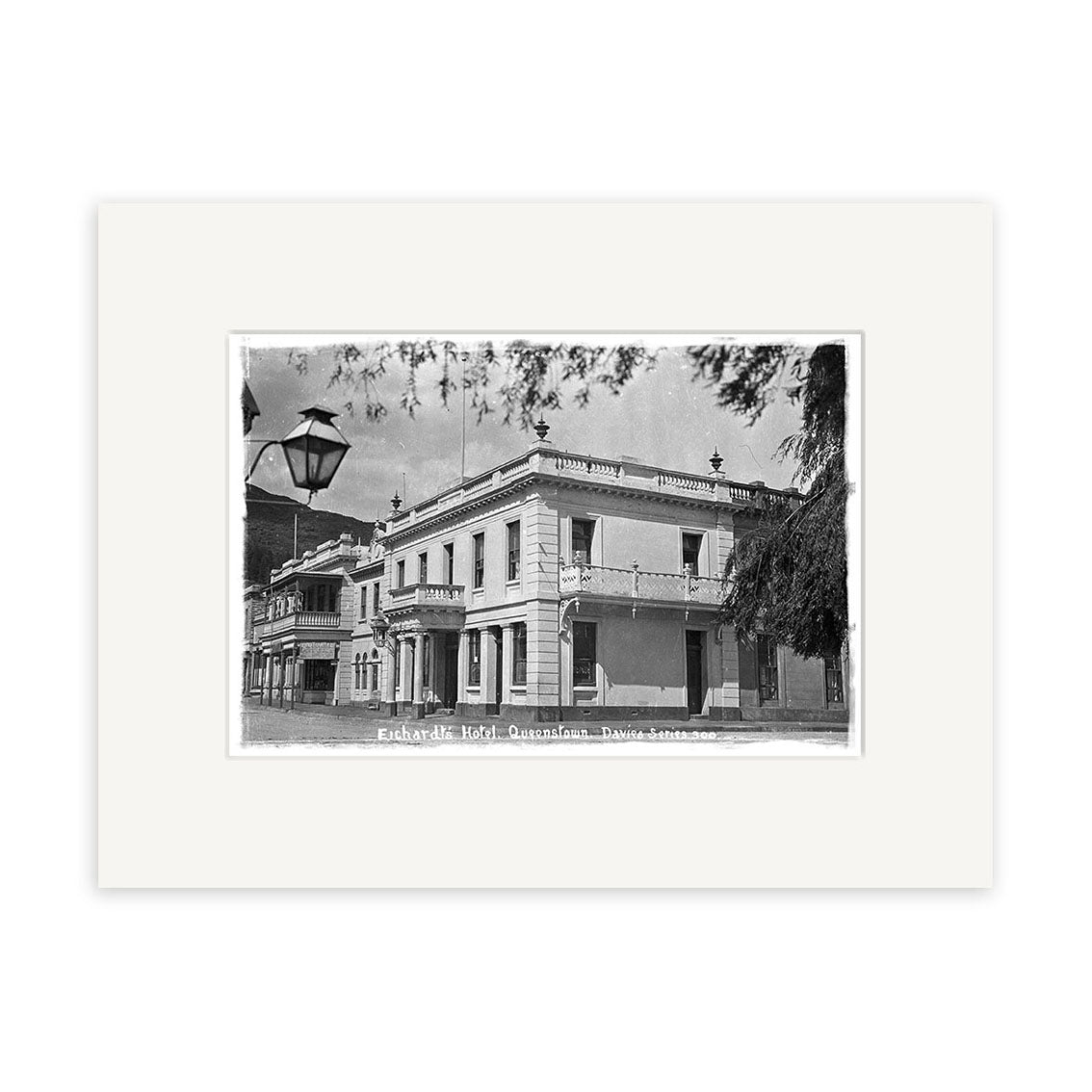 Black and white archival photo print of Eichardts Hotel, 1910 (HI118), a historic two-story building on a corner. Foreground includes streetlamp and tree branches. Labeled “Eucharda Hotel, Queensland, Parson’s Stores.”.