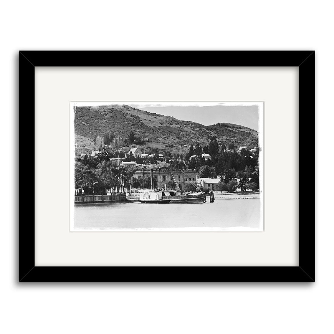 Archival black-and-white photo print, Queenstown 1910 - HI120, in a black frame shows a steamboat on a river before hillside houses and trees in historic New Zealand, with calm hills rising in the background.