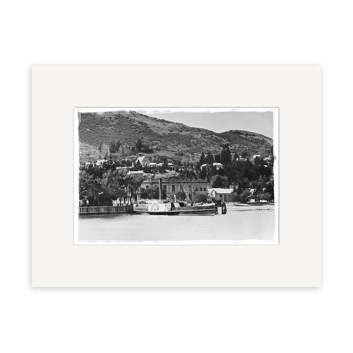 A black-and-white historical photographic print titled Queenstown 1910 - HI120 features a steamboat docked by a New Zealand waterfront, with houses, trees, and hills in the background under a clear sky. Matted with a white border.