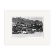 A black-and-white historical photographic print titled Queenstown 1910 - HI120 features a steamboat docked by a New Zealand waterfront, with houses, trees, and hills in the background under a clear sky. Matted with a white border.