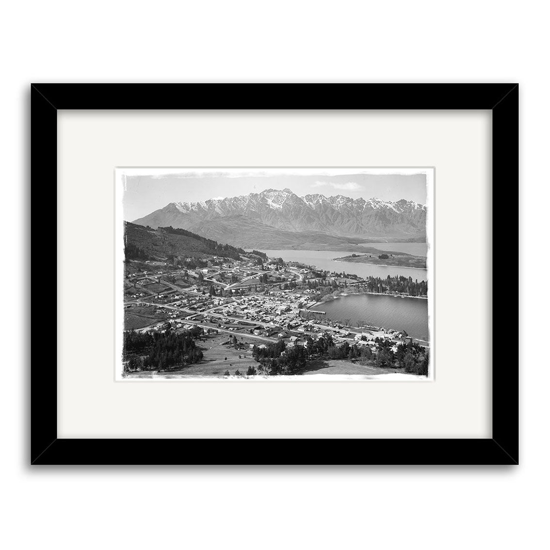 Black and white framed historical photographic print, Queenstown - HI121, shows a lakeside town with mountains and trees under a partly cloudy sky. Archival quality print. Made in New Zealand.