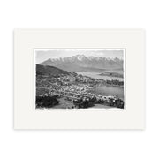 Black and white historical photo print Queenstown - HI121: a town beside a large lake with snow-capped mountains, grid streets, trees, and open land in New Zealand; mounted with a white mat.