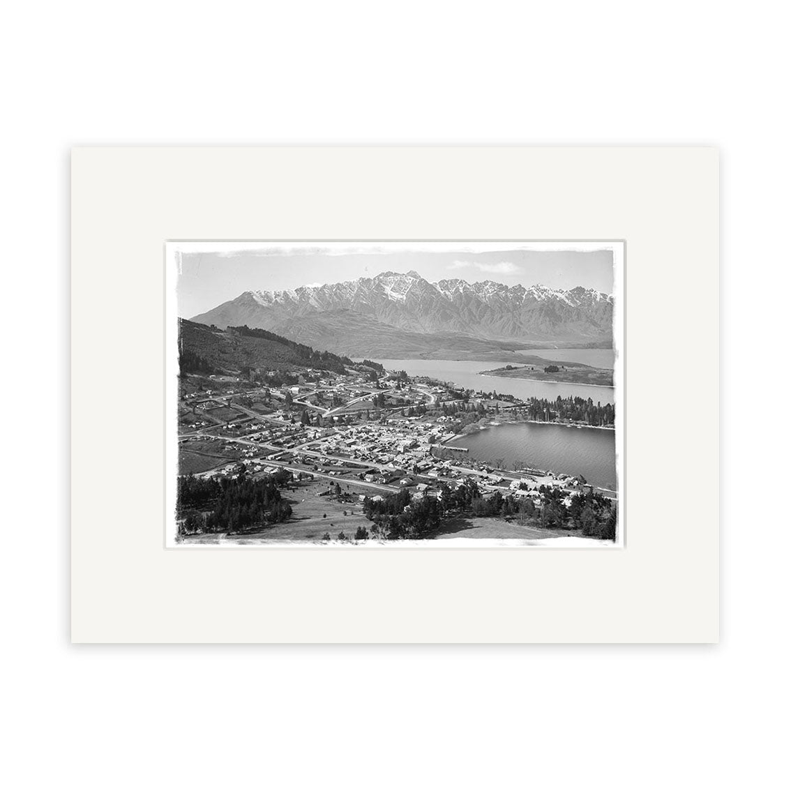 Black and white historical photo print Queenstown - HI121: a town beside a large lake with snow-capped mountains, grid streets, trees, and open land in New Zealand; mounted with a white mat.