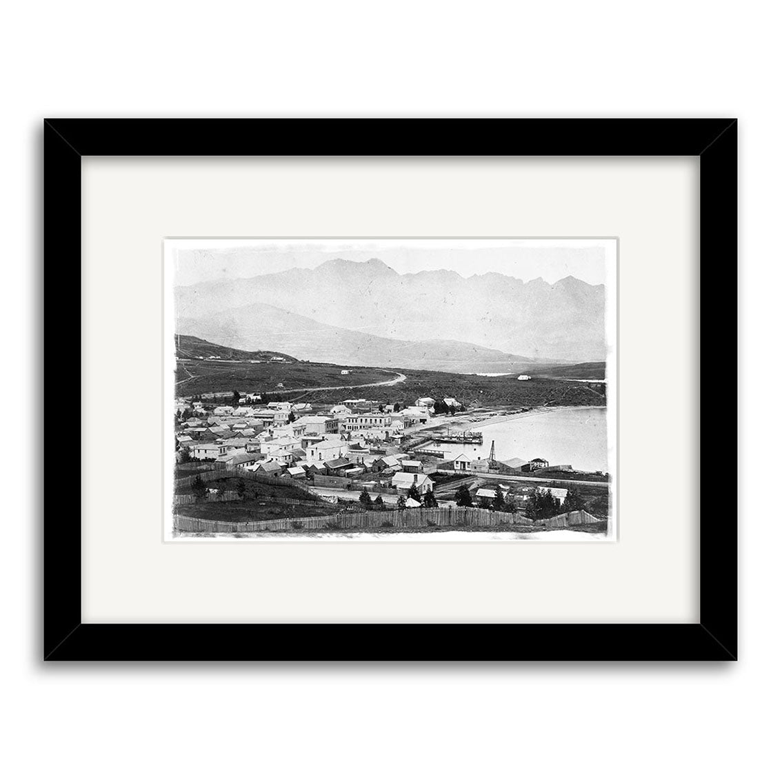 Black and white historical print, Queenstown 1880 - HI122: a small coastal town with clustered buildings, water to the right, and distant mountains. Printed on archival matte paper, framed with a white mat and black frame. Made in New Zealand.