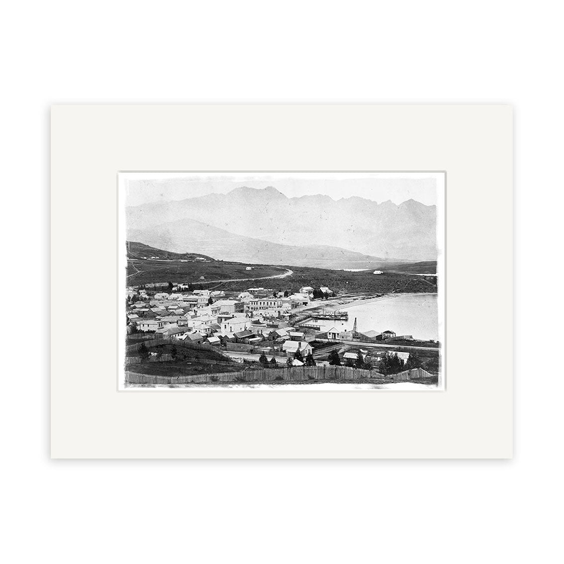 A historical black and white photo titled Queenstown 1880 - HI122, showing a small New Zealand town by the water with buildings along the shore and hills behind, printed on archival matte paper.