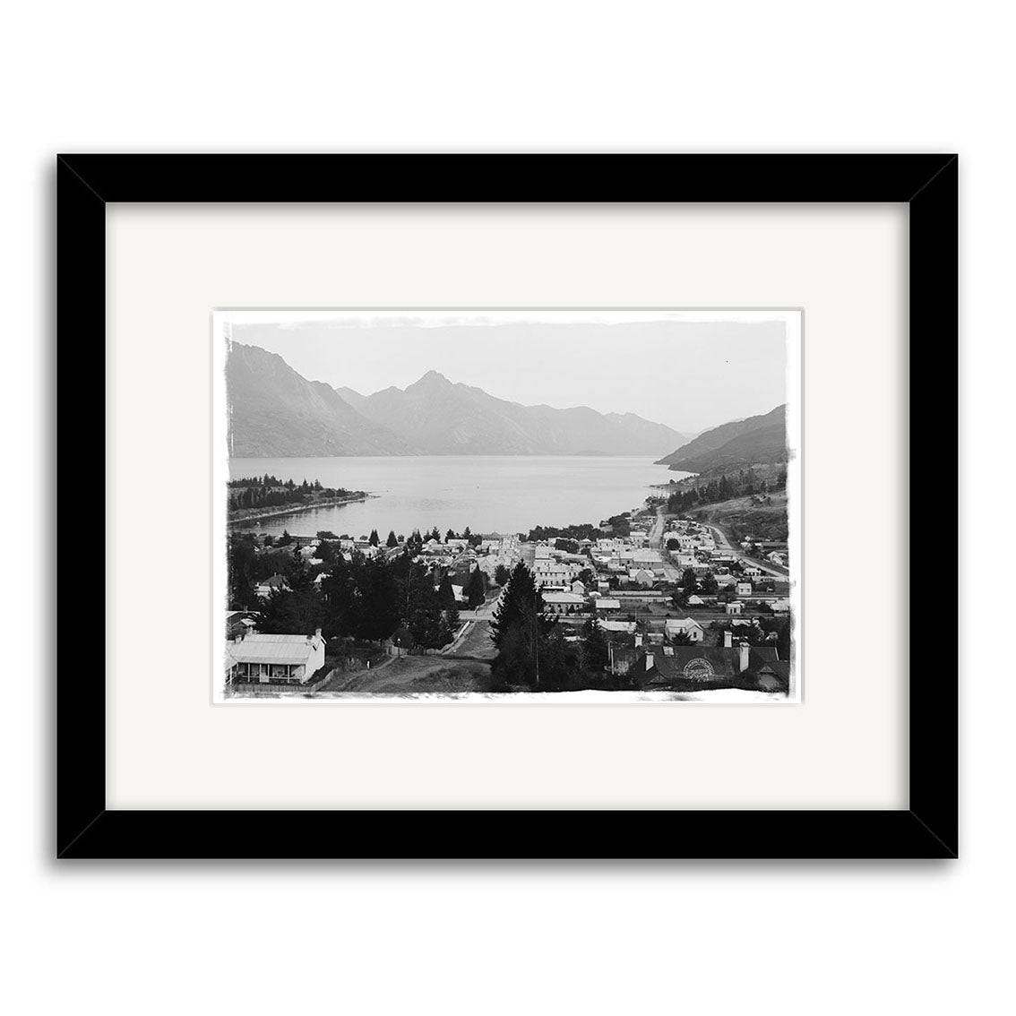 Black and white photograph Queenstown 1905 - HI123 of a lakeside New Zealand town with buildings, trees, and mountains, displayed in a black frame with a white mat on archival quality matte paper.