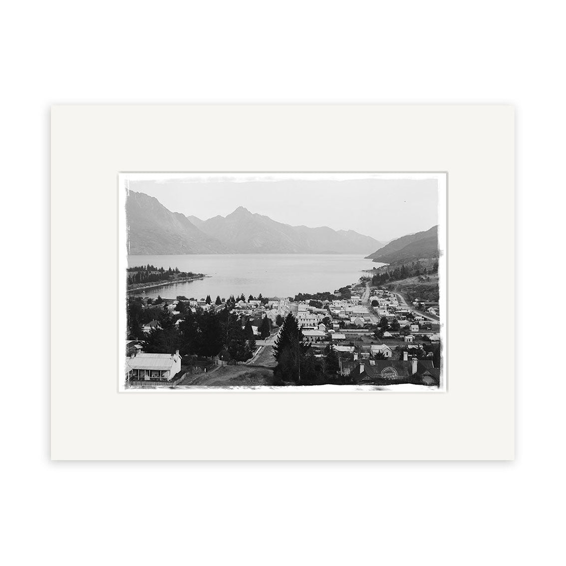 Black-and-white photo Queenstown 1905 - HI123: a calm New Zealand lakeside town with buildings, trees, and mountains. Historical photographic print, matted with a white border on archival-quality matte paper.