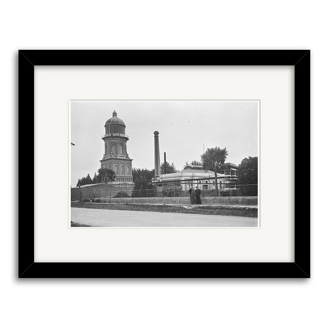 The Water Tower 1925 - HI124 is a black and white historical photo print of a domed building, smokestack, and structures in New Zealand with two people on a path. Presented in an archival-quality black frame with white mat.