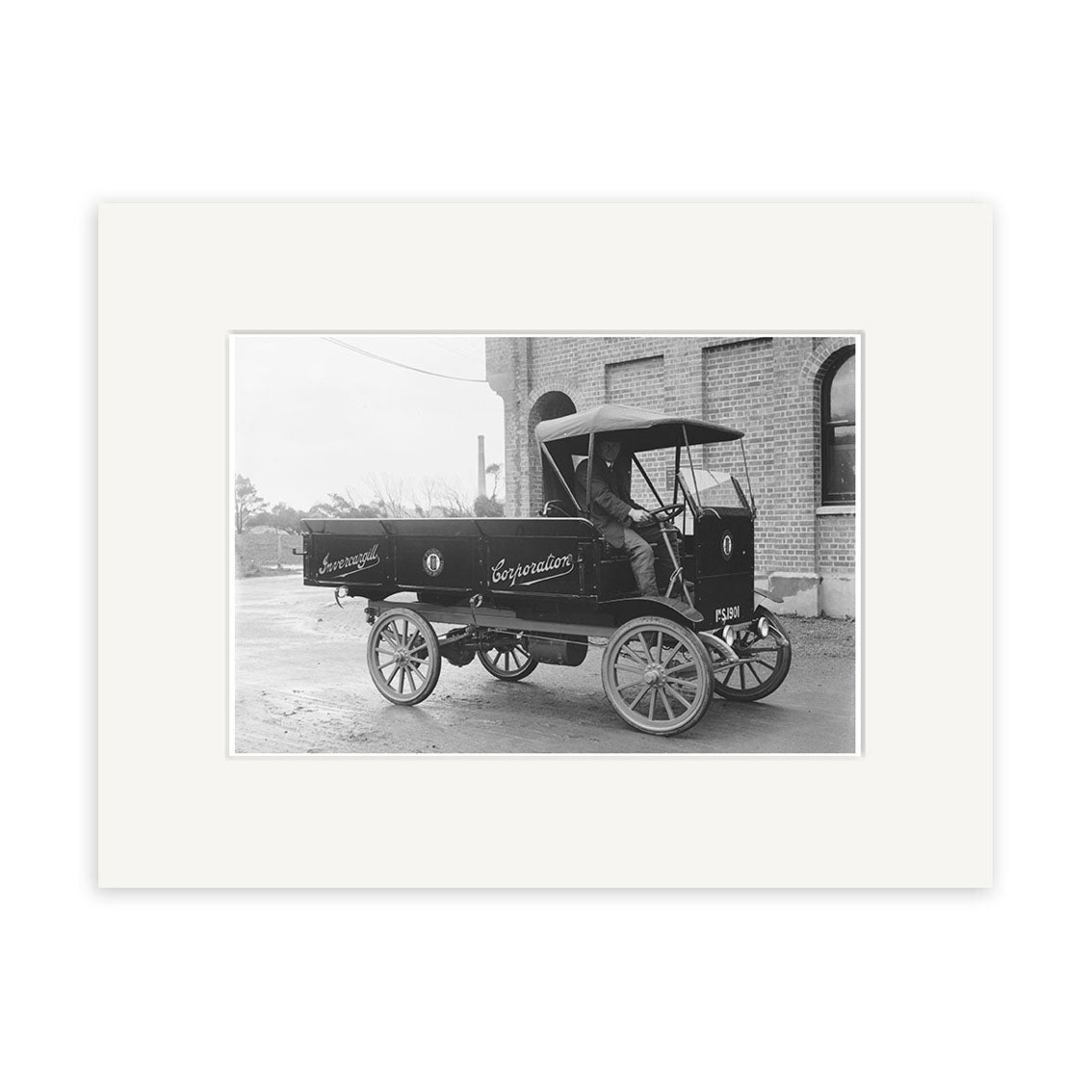 A black and white photo depicts the Electric Truck Invercargill 1920 - HI127 with wooden wheels and an open cabin, parked outside a brick building in New Zealand, driven by a visible driver.