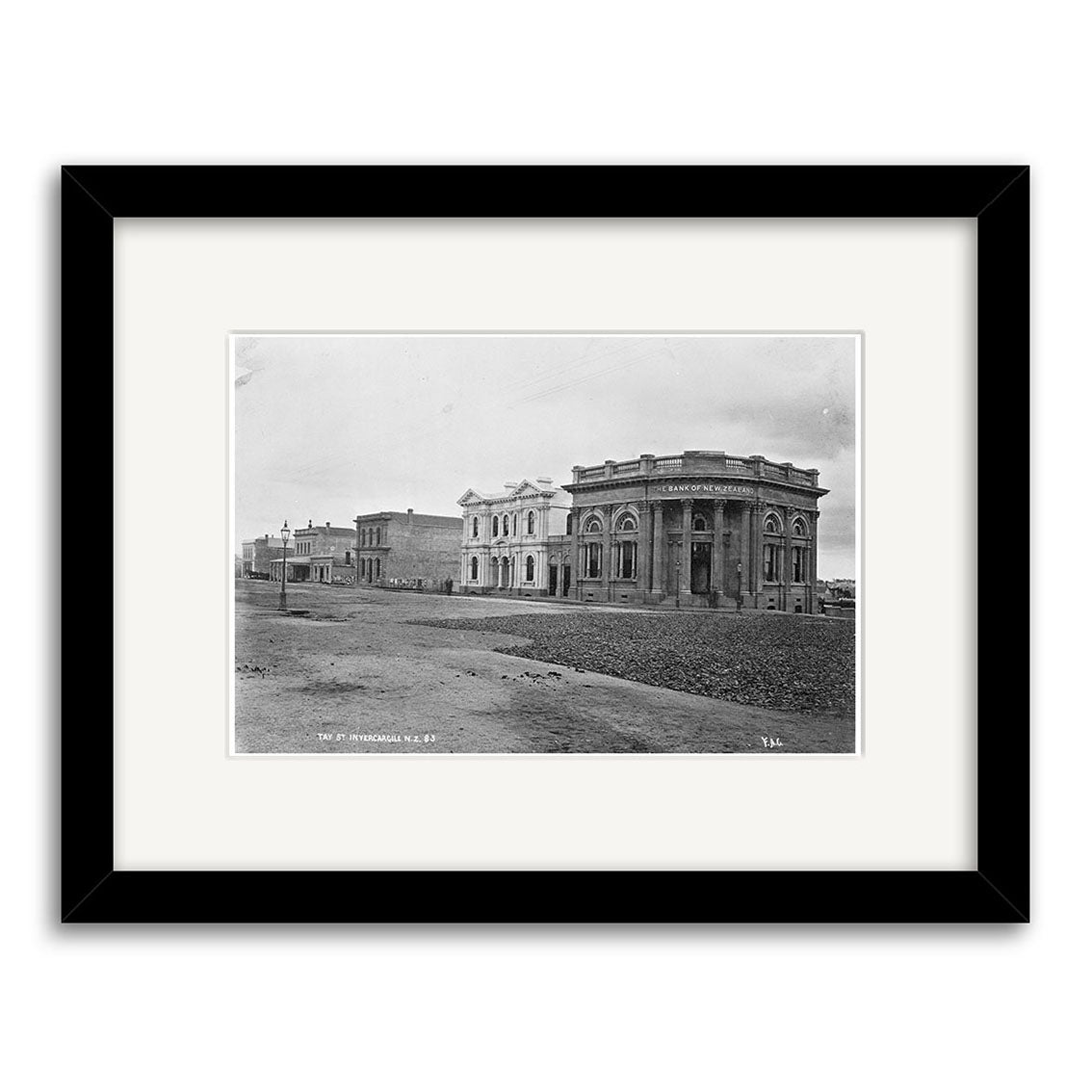 The Tay Street 1880 - HI129 is an archival black-and-white photo print of a mostly empty New Zealand street, featuring 19th-century stone buildings and a prominent corner bank. Framed in black with a white mat.