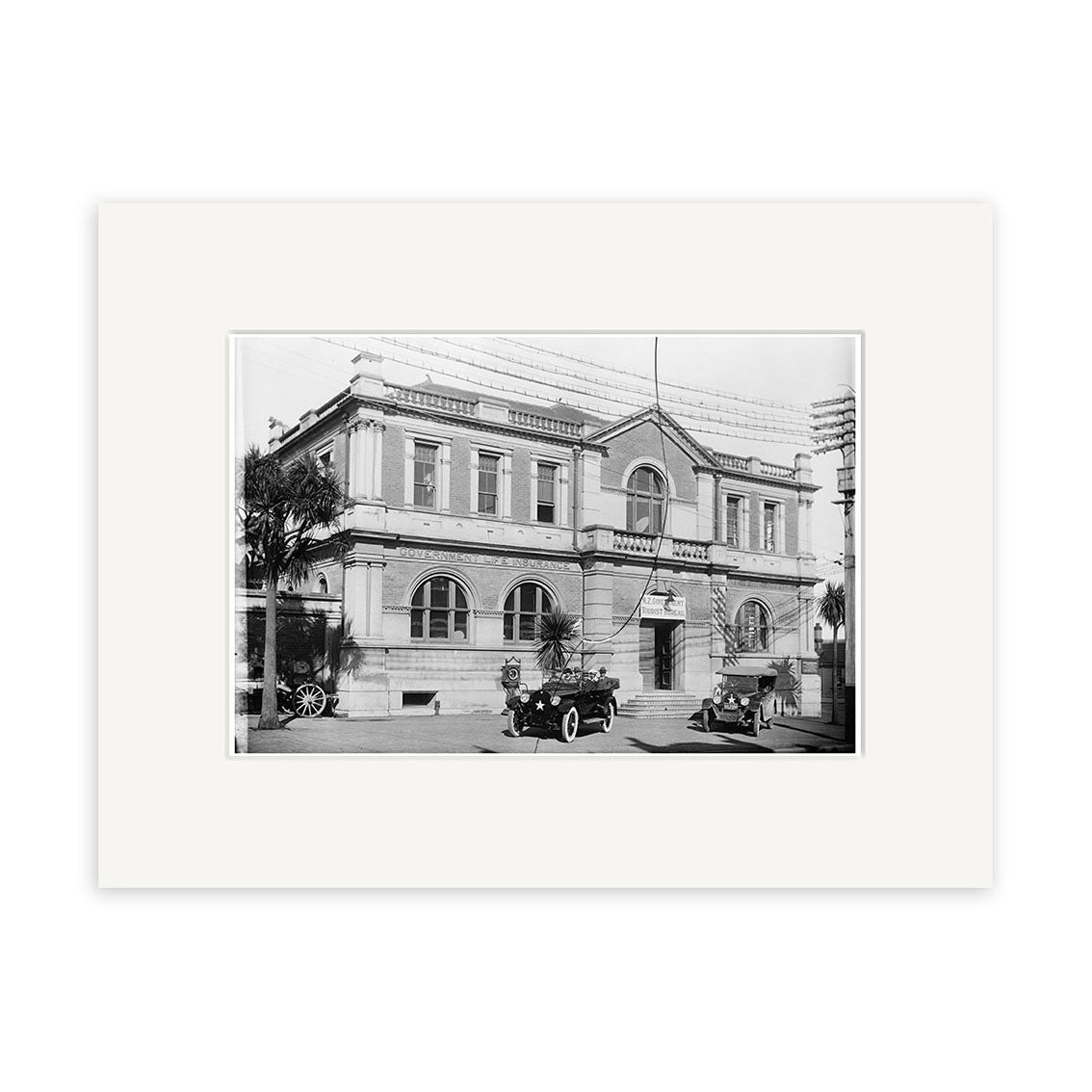 Black and white photo of a two-story classical building with arched windows, columns, and vintage cars out front. Palm trees line the side in this historical print: Govt Building 1922 - HI131, made in New Zealand.