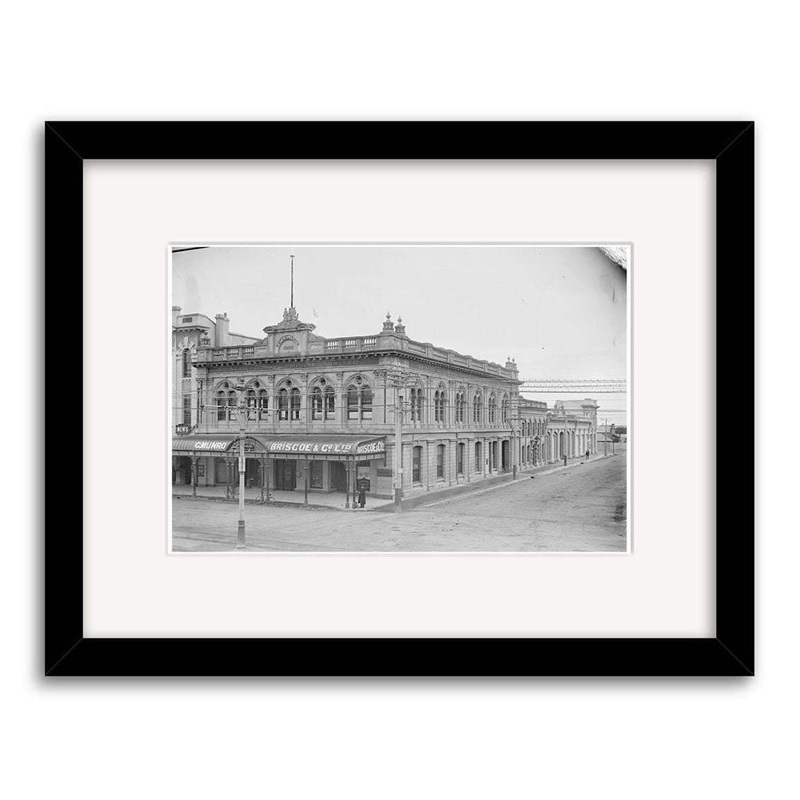 Black and white photo of Briscoe & Co Ltd 1890s - HI134, a two-story ornate corner building with arched windows, framed in black. Made in New Zealand on archival quality matte paper.