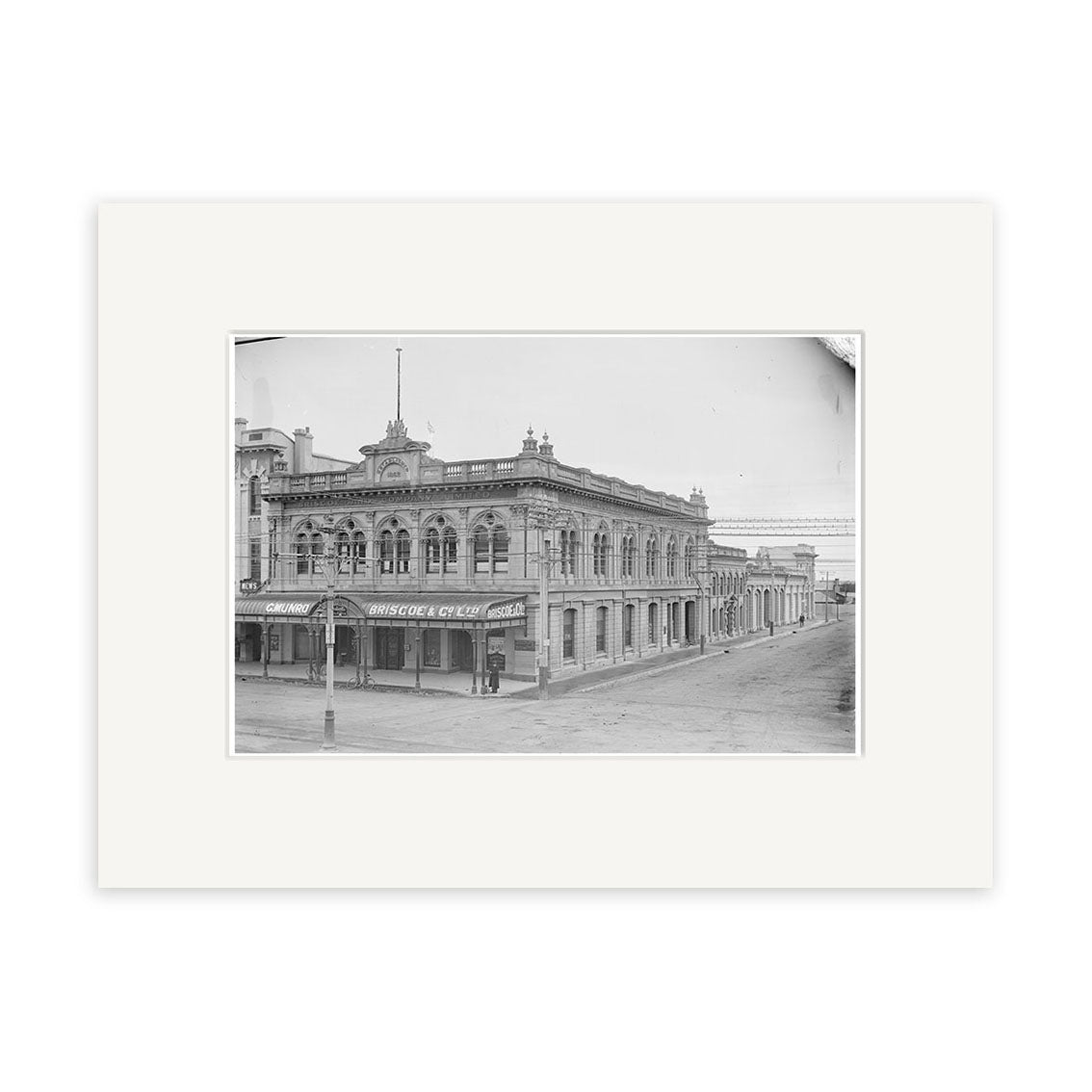 This archival quality matte print features a black and white photo of the two-story Briscoe & Co Ltd building from the 1890s (HI134), captured in New Zealand, with ornate details and a white mat border.