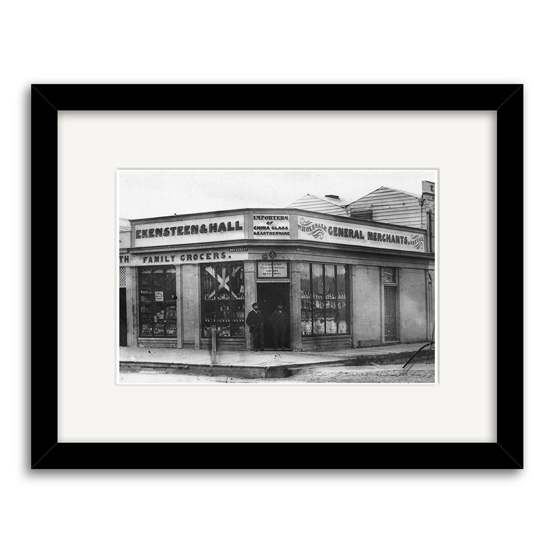 Black-and-white photo, Ekensteen & Halls 1880 – HI135: A person stands at the entrance of a family grocer’s shop, “Ekensteen & Hall,” with window advertisements. Printed on archival quality matte paper.