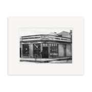 Black-and-white archival matte print of Ekensteen & Halls 1880 (HI135), showing a corner store with GROCER and GENERAL MERCHANT signs. Two people stand at the entrance.
