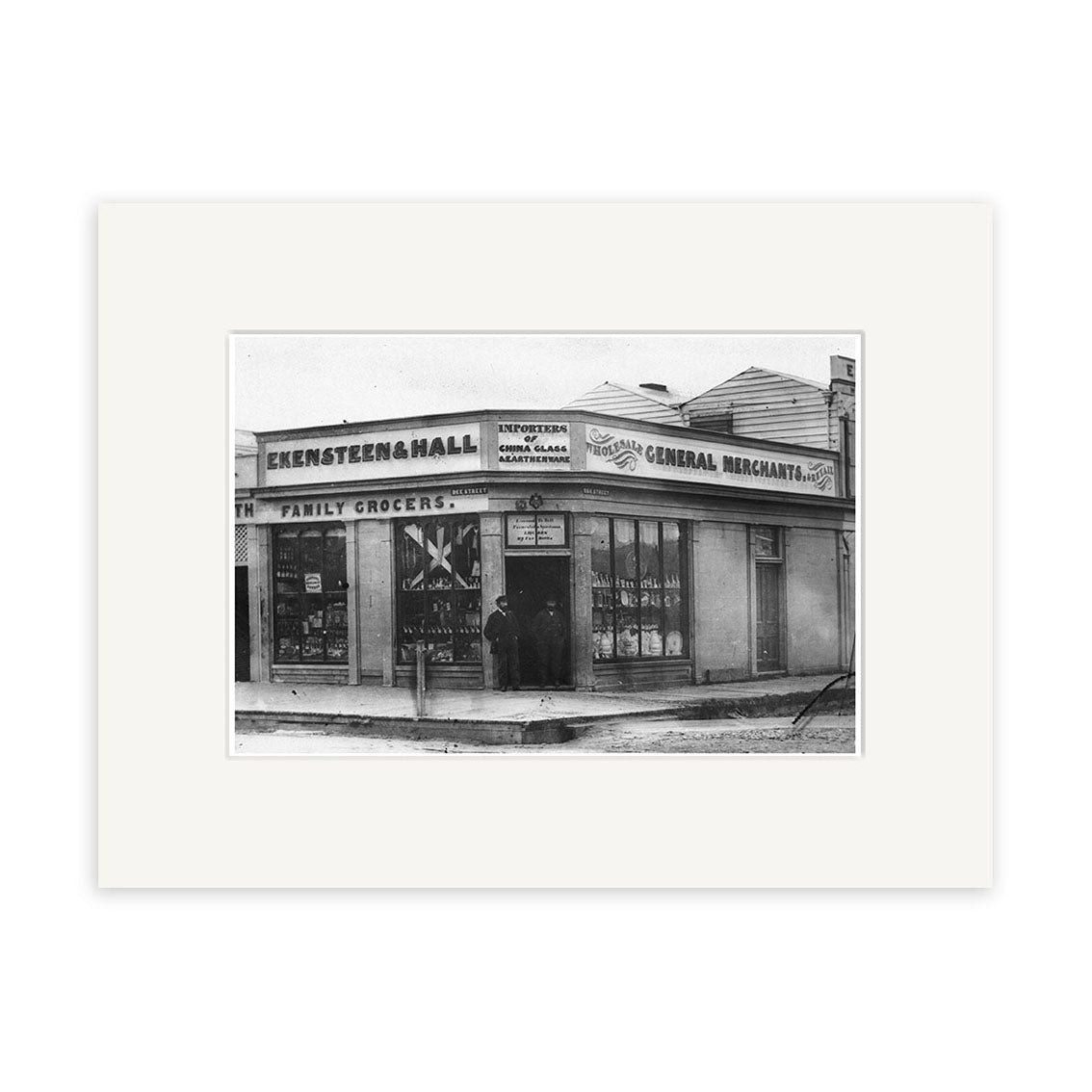 Black-and-white archival matte print of Ekensteen & Halls 1880 (HI135), showing a corner store with GROCER and GENERAL MERCHANT signs. Two people stand at the entrance.