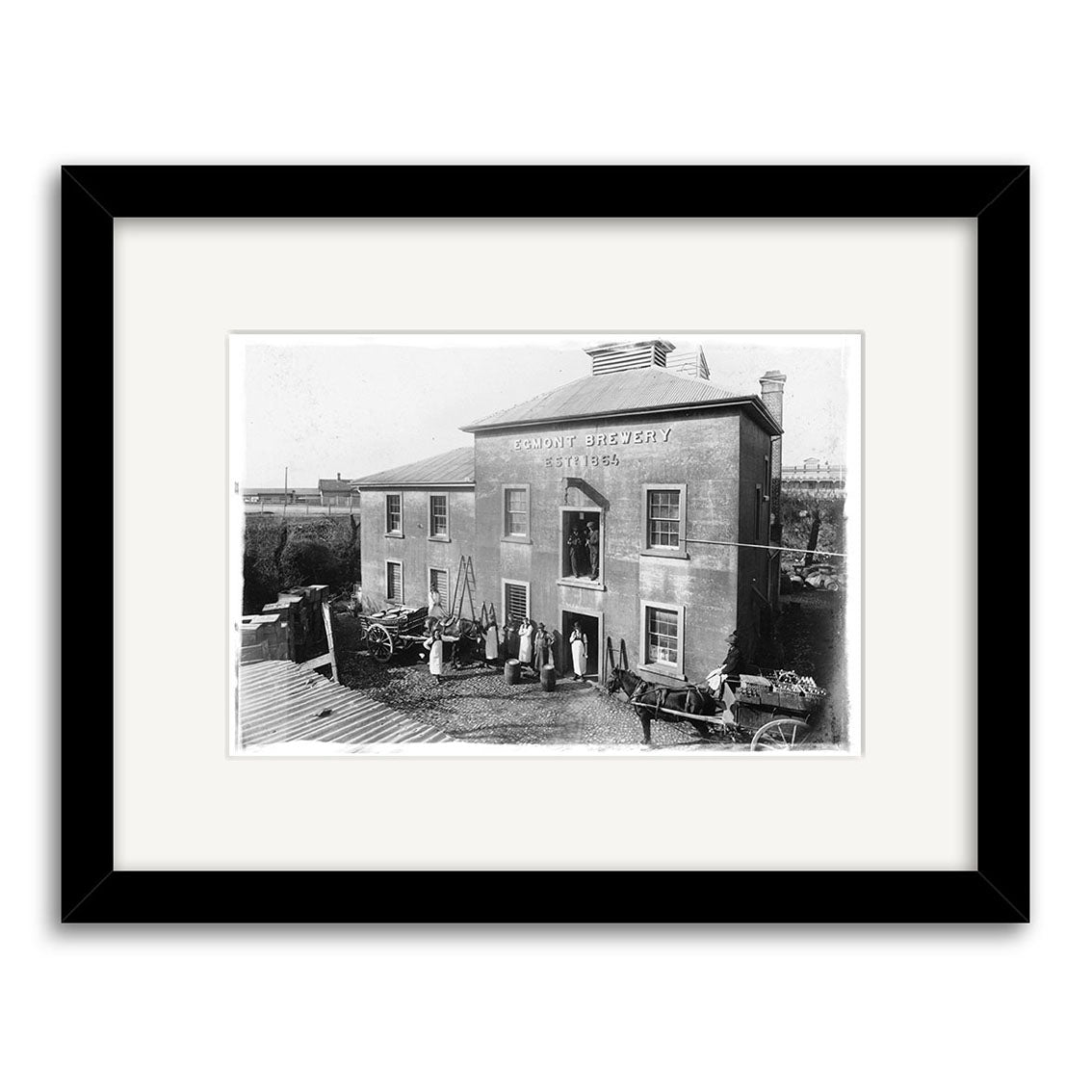 Black-and-white historical print of Egmont Brewery 1906 - HI138 in New Zealand, showing people with horse-drawn wagons and tools. Presented with archival quality matting and a black frame.