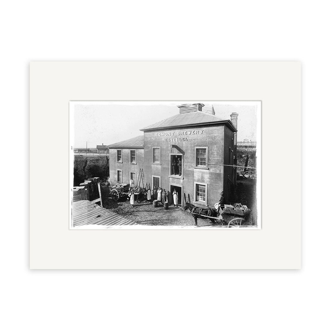 Archival-quality black and white historical photo print of Egmont Brewery 1906 (HI138), showing a two-story building in New Zealand with people, carts, gardening tools, a fence, and trees in the background.