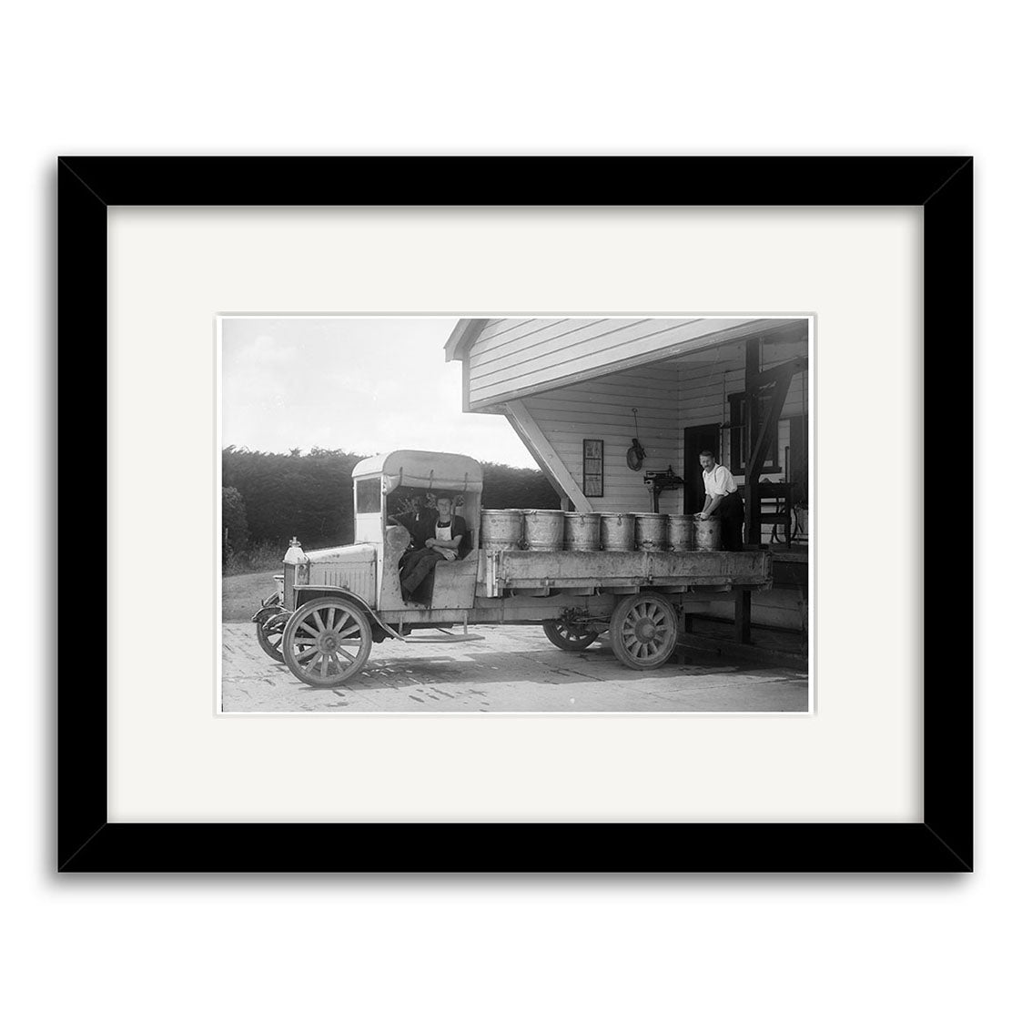 Milk Truck Taranaki 1918 - HI141 is a framed, matted historical print showing an early 20th-century New Zealand truck with milk cans at a dock. Two people sit in the cab, one stands nearby holding a can.