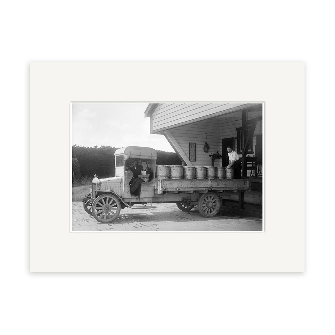 The archival-quality black-and-white photo Milk Truck Taranaki 1918 - HI141 shows an old-fashioned truck with milk cans parked by a building in New Zealand, two men in the cab, and a third on the loading dock.